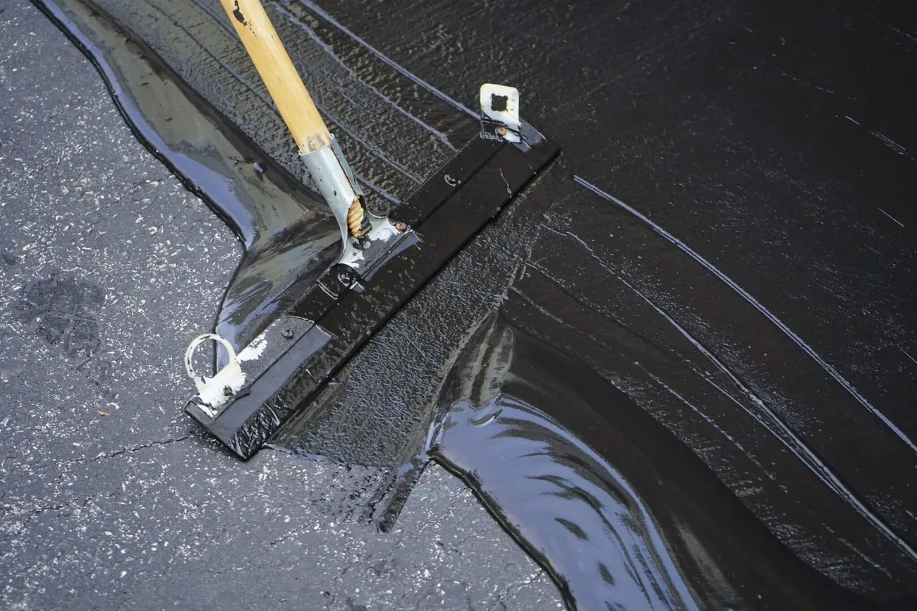 A squeegee spreading black sealcoat over a prepped asphalt driveway surface.